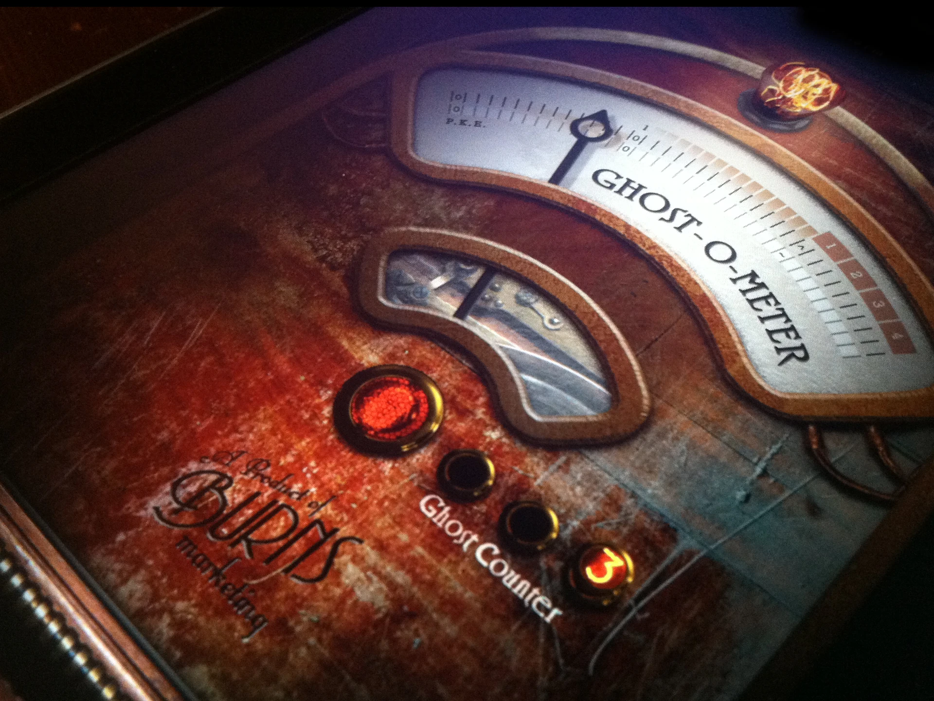 A close-up of the Ghost-O-Meter interface — a brass P.K.E. gauge, a glowing red button, and a “Ghost Counter,” all on a scratched, aged background marked “a product of Burns marketing.”