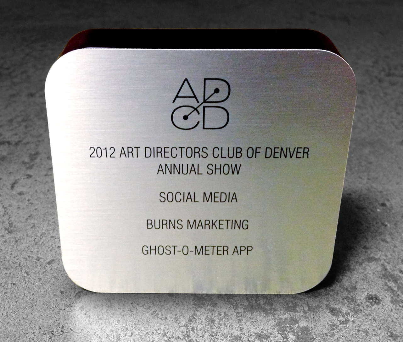 The Art Directors Club of Denver award — a brushed-metal trophy engraved “2012 ADCD Annual Show · Social Media · Burns Marketing · Ghost-O-Meter App.”