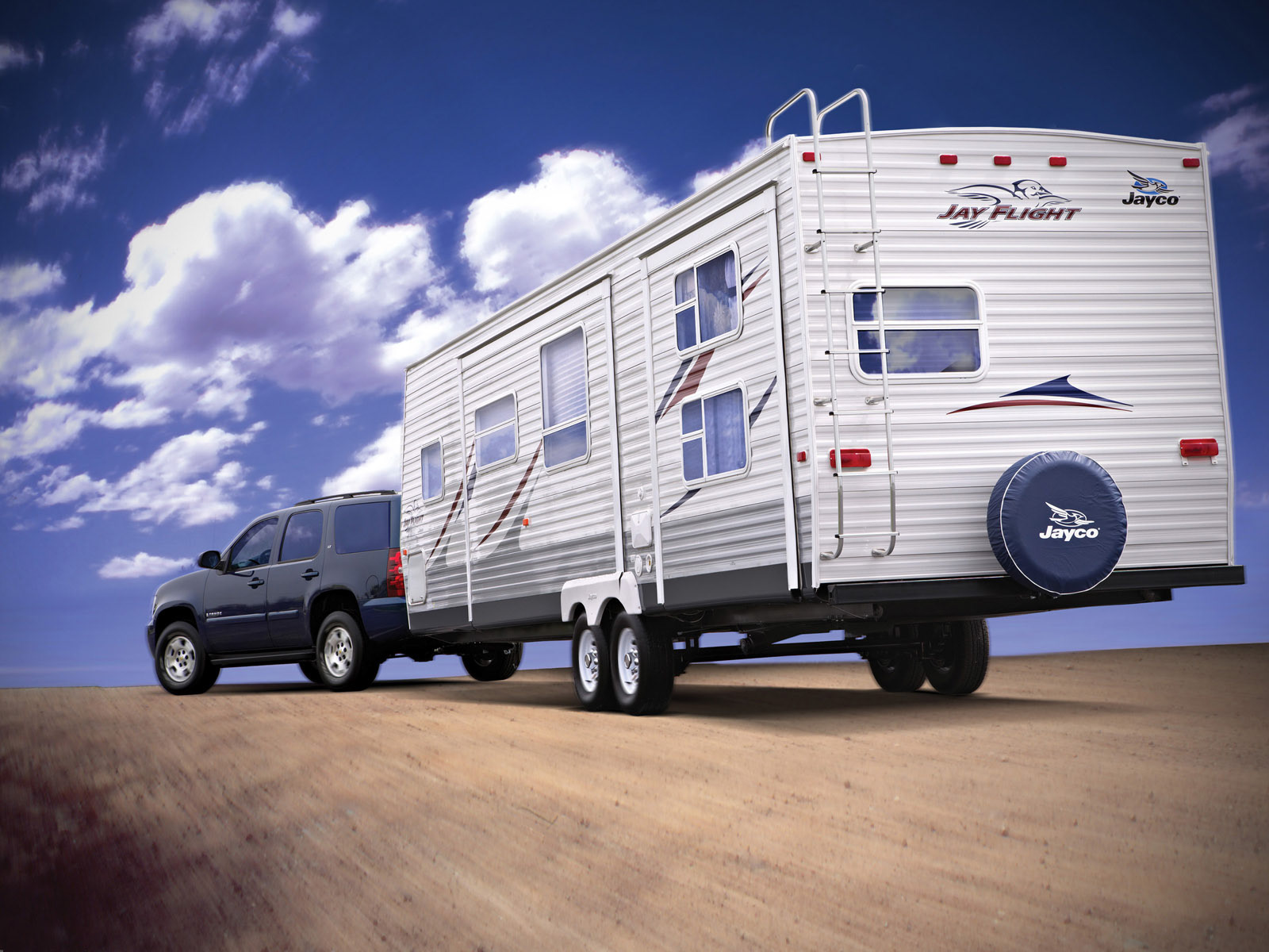 A Jayco “Jay Flight” travel trailer towed by an SUV across an open desert road under a dramatic sky — art-directed product photography.