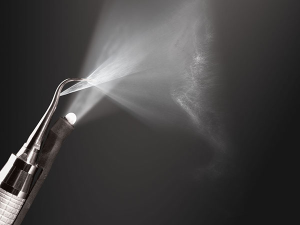 A dramatic black-and-white product photo of a Zila dental instrument emitting a fine spray, lit to feel precise and high-end.